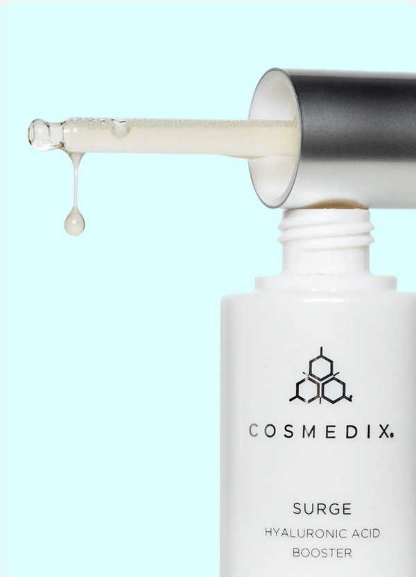 Close-up of a cosmetic dropper dispensing a droplet of serum above a white Cosmedix Surge Hyaluronic Acid Booster bottle against a light blue background.